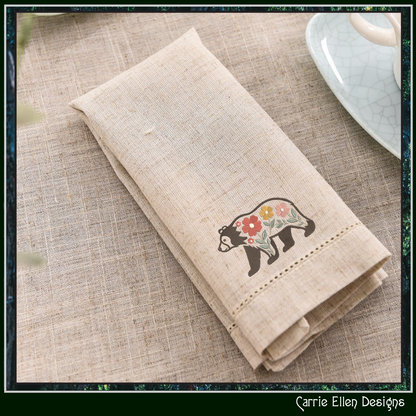 Folded beige napkin with a bear design embroidered on a textured surface, featuring 'Carrie Ellen Designs' branding.